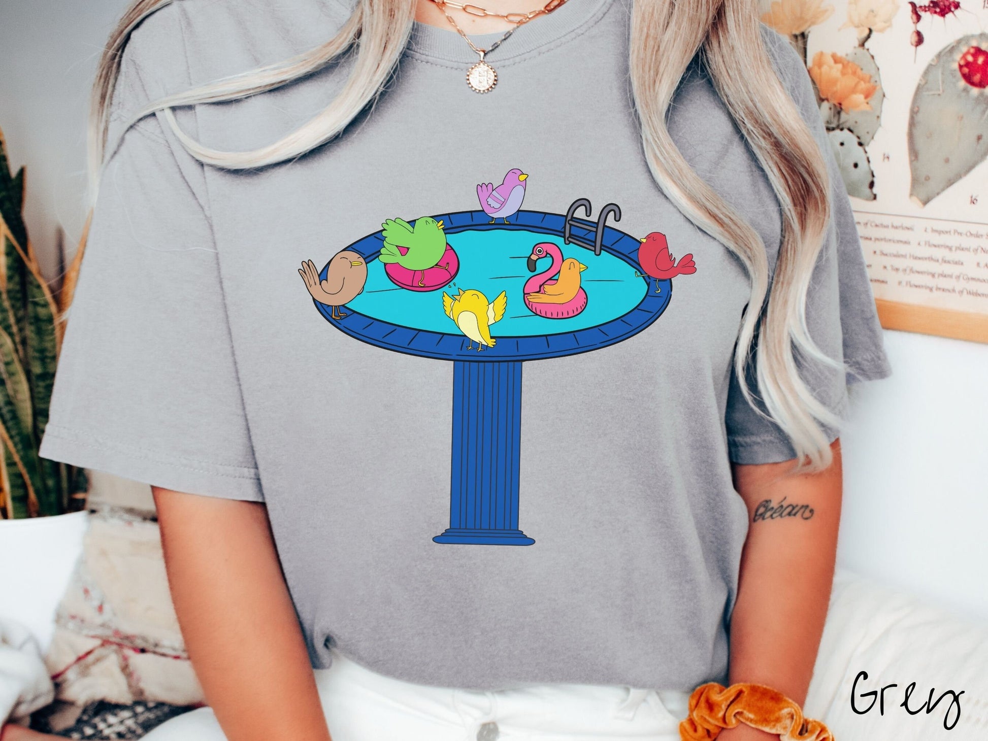 A woman wearing a cute, vintage grey colored Comfort Colors shirt with a blue birdbath with a pool ladder filled with crystal clear blue water. In the water are colorful birds floating on colorful pool inflatables and enjoying the sun.