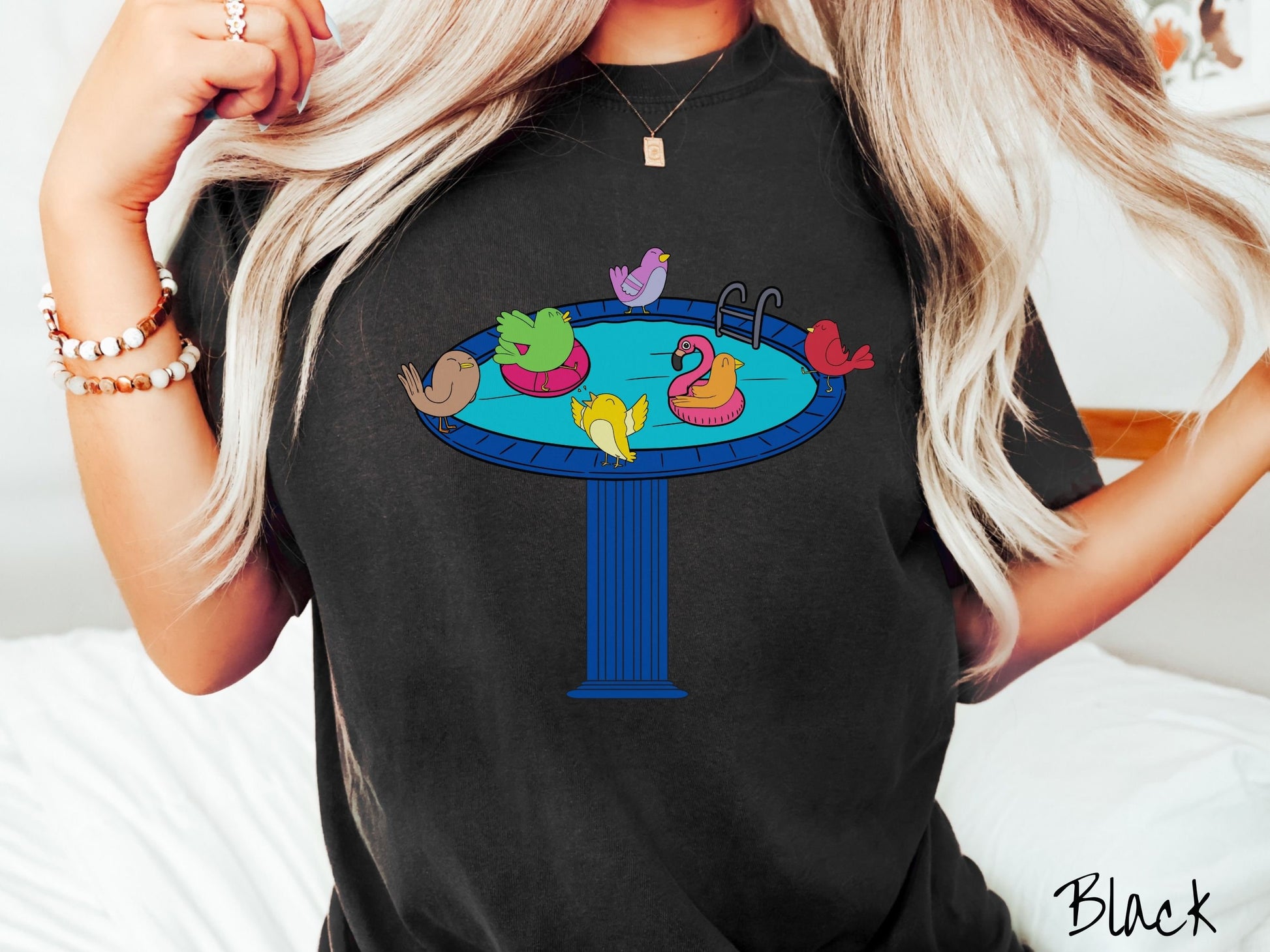 A woman wearing a cute, vintage black colored Comfort Colors shirt with a blue birdbath with a pool ladder filled with crystal clear blue water. In the water are colorful birds floating on colorful pool inflatables and enjoying the sun.