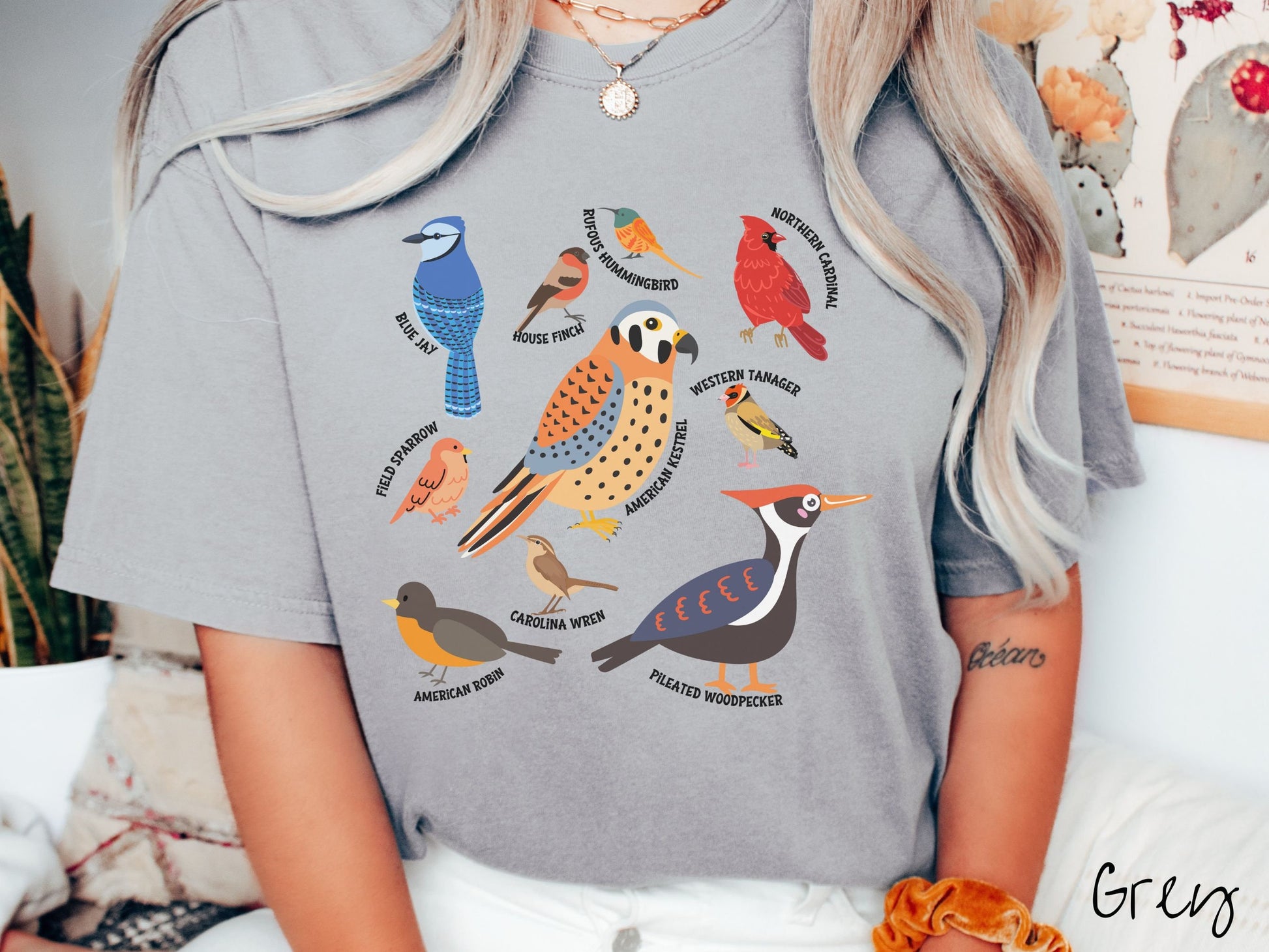 A woman wearing a cute, vintage grey colored Comfort Colors shirt with pictures of nine different bird species and their names listed across the front. Some examples are the Blue Jay, House Finch, Western Tanager, and Carolina Wren.