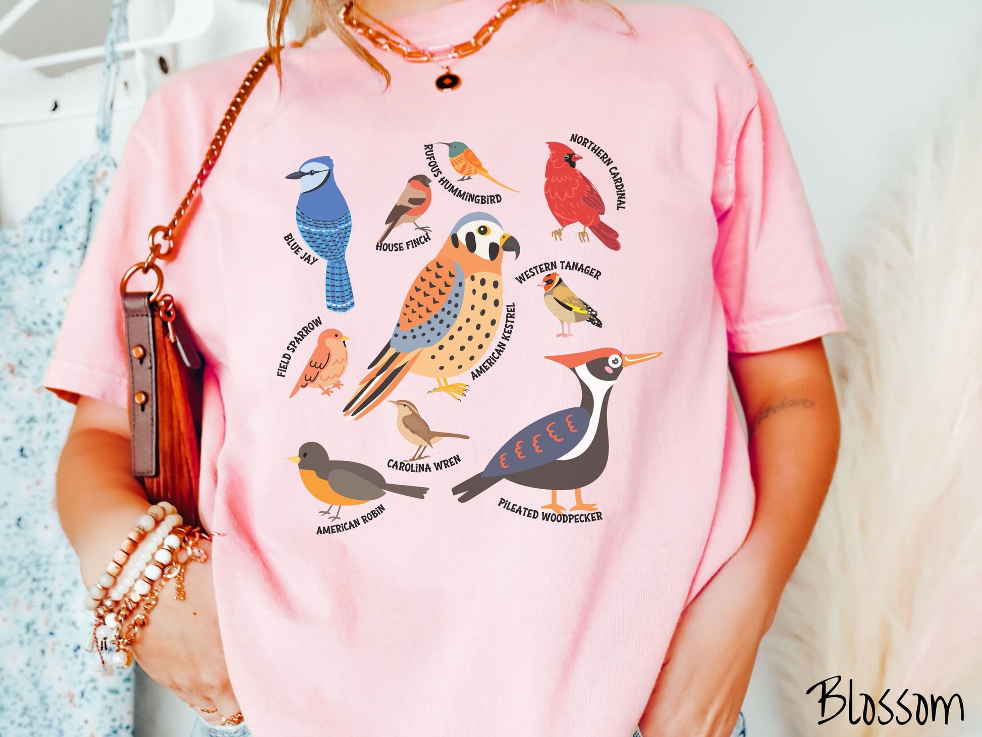 A woman wearing a cute, vintage blossom colored Comfort Colors shirt with pictures of nine different bird species and their names listed across the front. Some examples are the Blue Jay, House Finch, Western Tanager, and Carolina Wren.