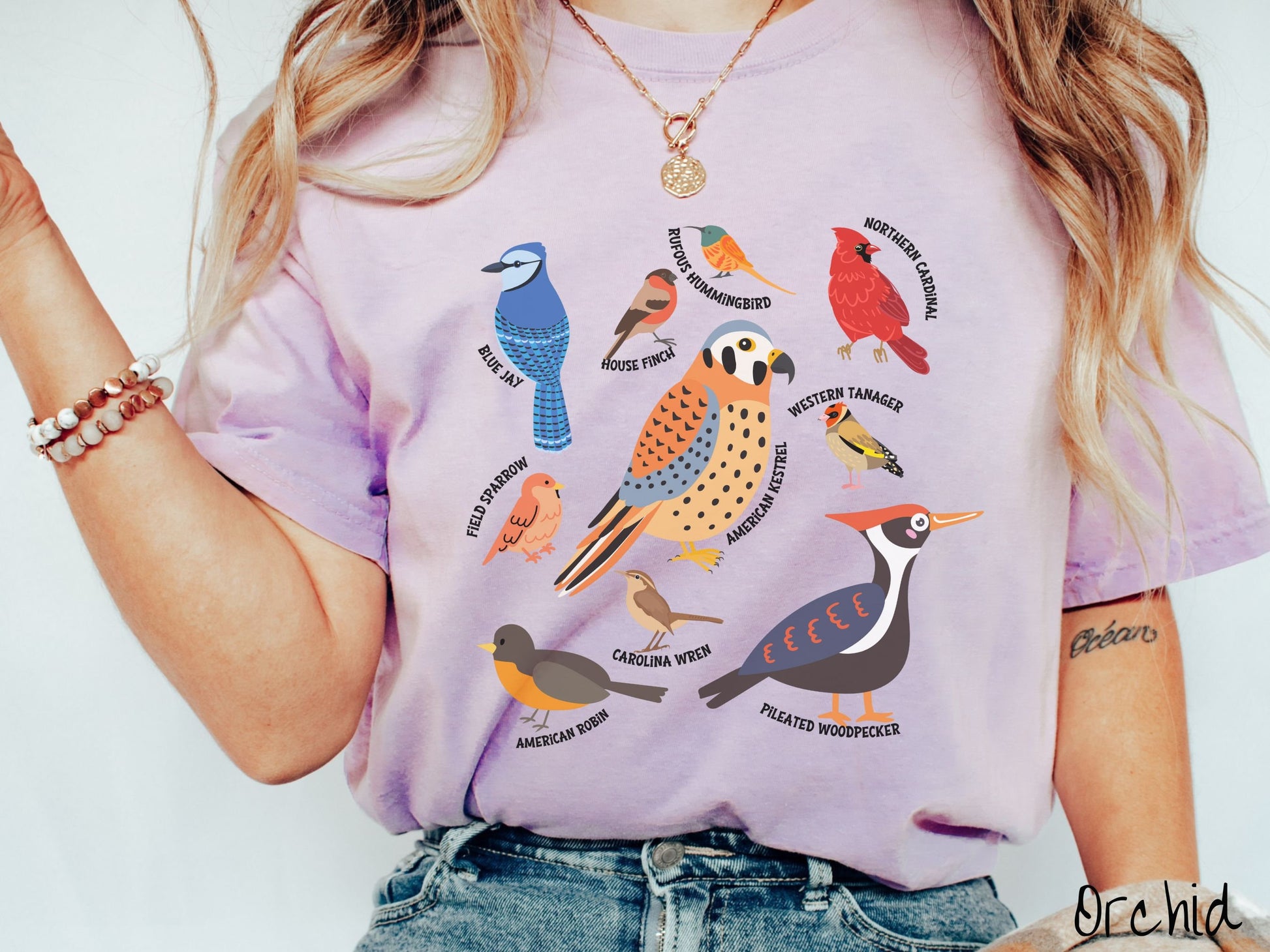 A woman wearing a cute, vintage orchid colored Comfort Colors shirt with pictures of nine different bird species and their names listed across the front. Some examples are the Blue Jay, House Finch, Western Tanager, and Carolina Wren.