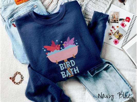 A woman wearing a cute, vintage navy blue colored comfy sweatshirt with a pink bathtub with a red bird and purple bird enjoying a bubble bath together. Below the bathtub are the words Bird Bath in light blue, uppercase font.