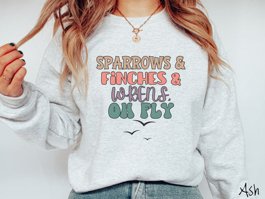 A woman wearing a cute, vintage ash colored comfy sweatshirt with the text Sparrows & Finches & Wrens, Oh Fly in brown, pink, purple, and green font. Below that are three black silhouettes of birds flying through the air.