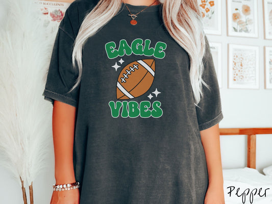 A woman wearing a cute pepper colored t-shirt with green text saying Eagle Vibes. There is a brown football with white twinkling stars around it between the text.