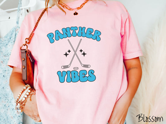 A woman wearing a cute blossom colored t-shirt with blue text saying Panther Vibes. There are two black outlined hockey sticks and a hockey puck with black twinkling stars around them between the text.