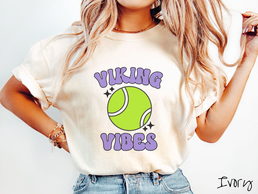 A woman wearing a cute ivory colored shirt with purple text saying Viking Vibes. There is a neon tennis ball with white stripes and black twinkling stars around it between the text.