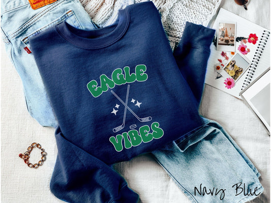 A cute navy blue colored sweatshirt with green text saying Eagle Vibes. There are two white outlined hockey sticks and a hockey puck with white twinkling stars around them between the text.