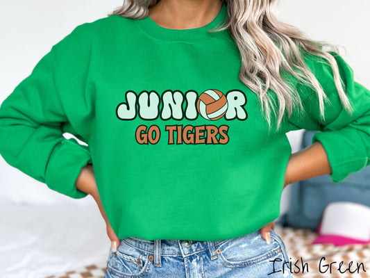 A woman wearing a cute Irish green colored sweatshirt with the word Junior in black and white font, the O in Junior is an orange and white volleyball. Below Junior are the words in orange and black font Go Tigers.