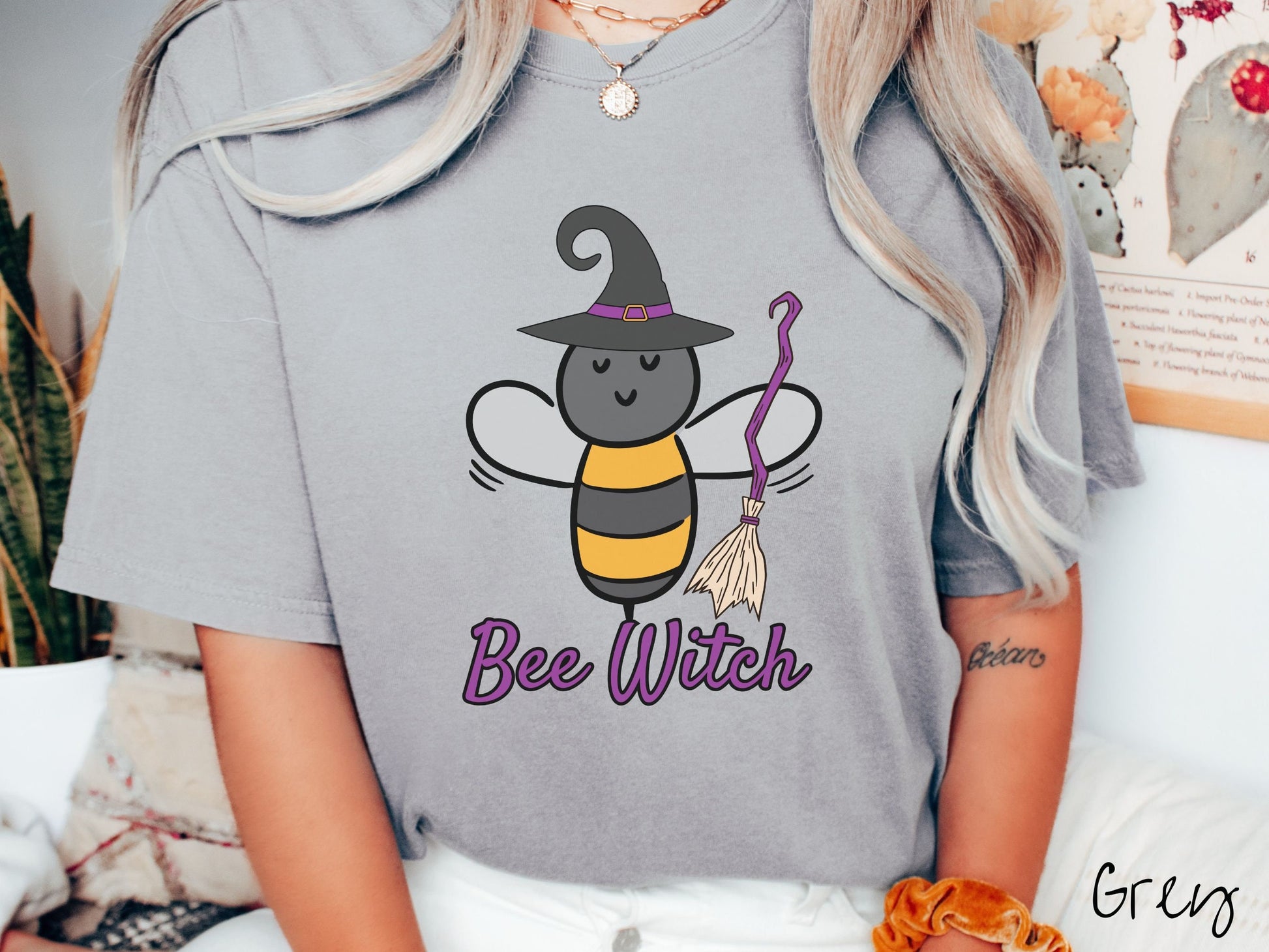A woman wearing a cute, vintage grey colored Comfort Colors t-shirt with the text Bee Witch in purple, cursive font. Above that is a black and yellow honey bee holding a purple, straw broom and wearing a black witch hat with a purple hat band.
