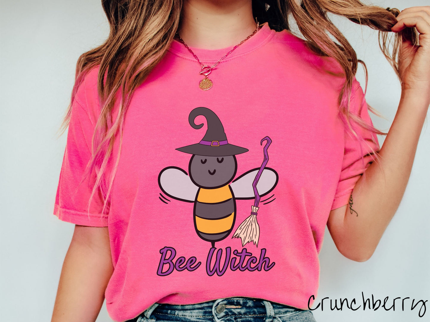 A woman wearing a cute, vintage crunchberry colored Comfort Colors t-shirt with the text Bee Witch in purple, cursive font. Above that is a black and yellow honey bee holding a purple, straw broom and wearing a black witch hat with a purple hat band.