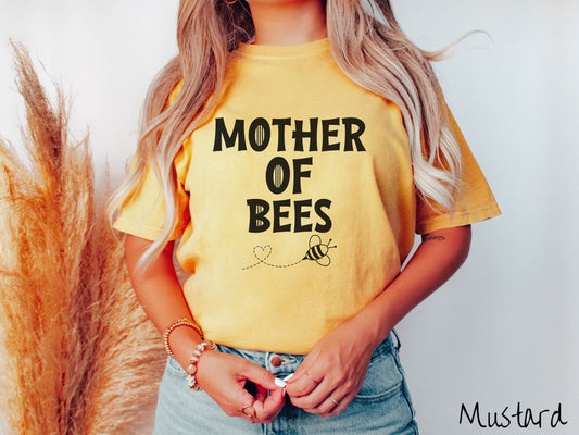 A woman wearing a cute, vintage mustard colored Comfort Colors t-shirt with the text Mother of Bees in black, uppercase font. Below that is a black outlined bee buzzing around, the trail it leaves has created a heart shape in the air.