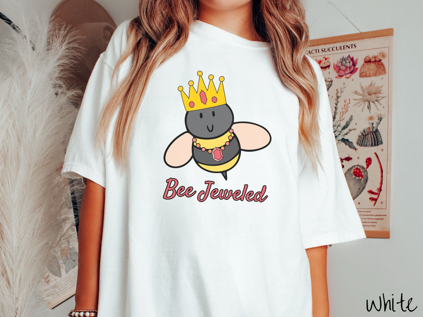 A woman wearing a cute, vintage white colored Comfort Colors T-shirt with the text Bee Jeweled in pink, cursive font. Above that is a black and yellow honey bee wearing a red, bejeweled golden crown, and a golden necklace with a large, red jewel.