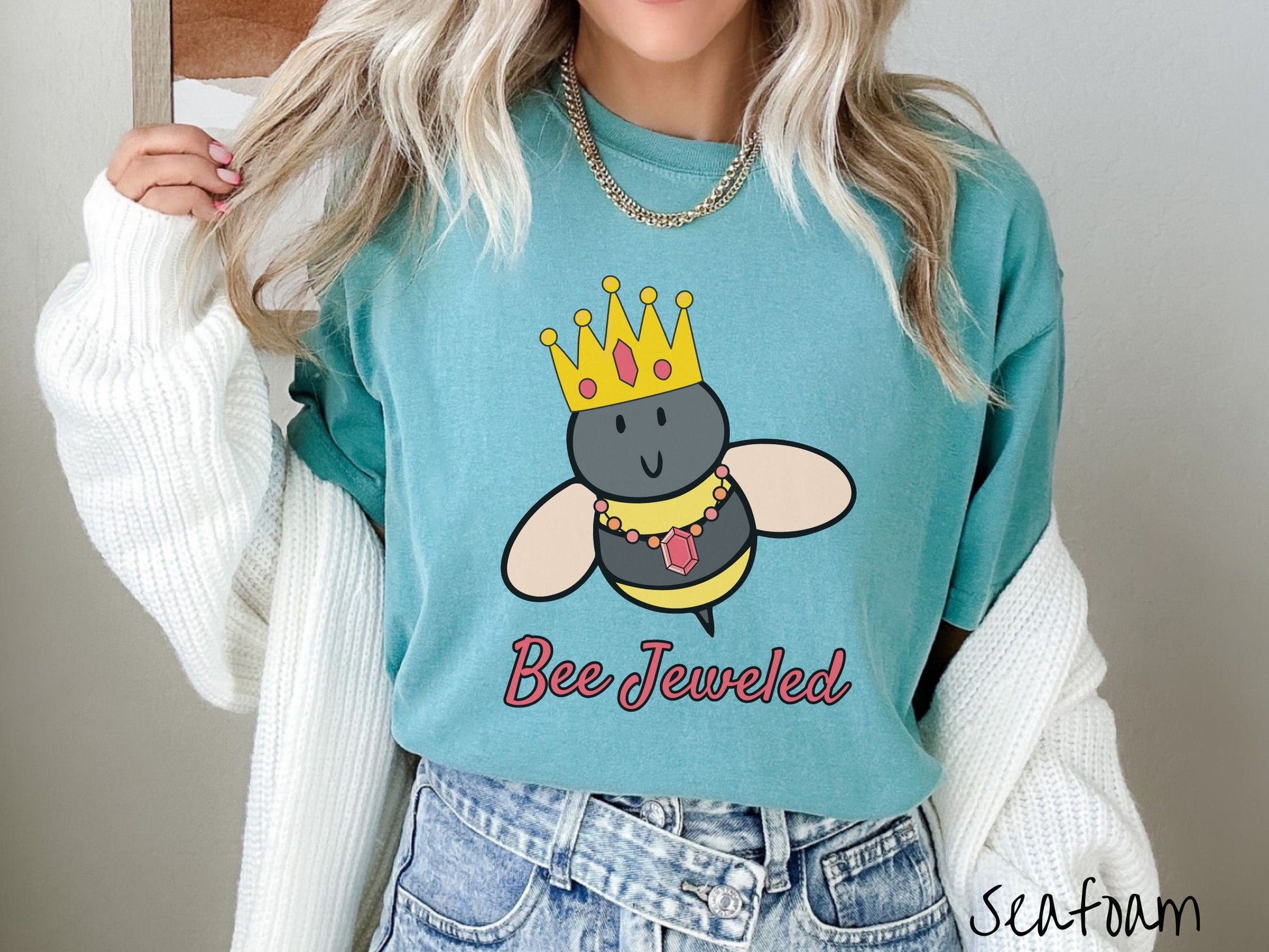 A woman wearing a cute, vintage seafoam colored Comfort Colors T-shirt with the text Bee Jeweled in pink, cursive font. Above that is a black and yellow honey bee wearing a red, bejeweled golden crown, and a golden necklace with a large, red jewel.