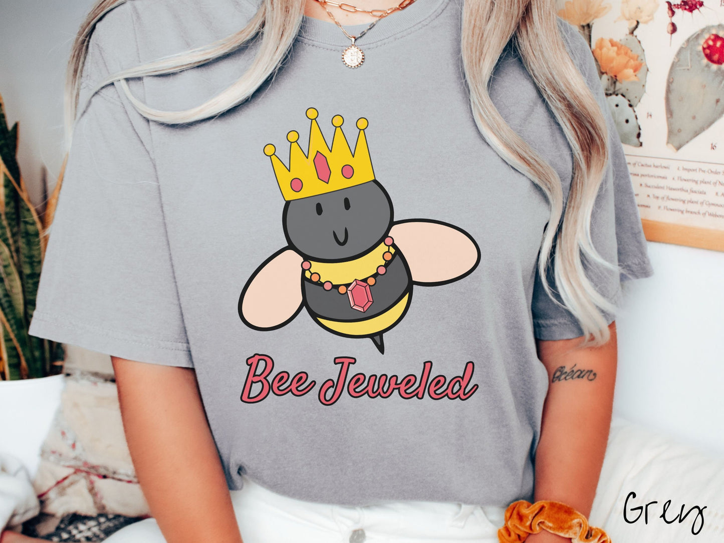 A woman wearing a cute, vintage grey colored Comfort Colors T-shirt with the text Bee Jeweled in pink, cursive font. Above that is a black and yellow honey bee wearing a red, bejeweled golden crown, and a golden necklace with a large, red jewel.