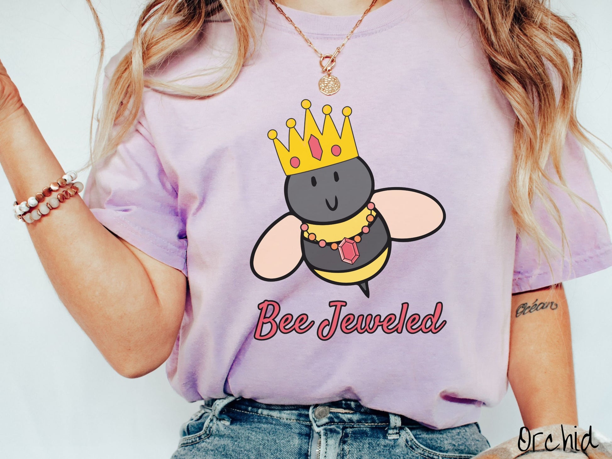 A woman wearing a cute, vintage orchid colored Comfort Colors T-shirt with the text Bee Jeweled in pink, cursive font. Above that is a black and yellow honey bee wearing a red, bejeweled golden crown, and a golden necklace with a large, red jewel.