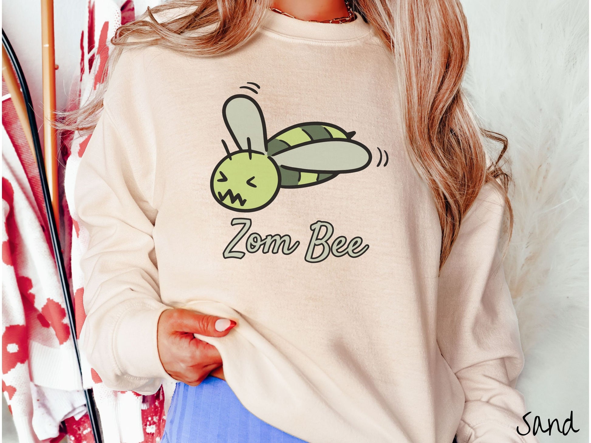 A woman wearing a cute, vintage sand colored comfy sweatshirt with the text Zom Bee in light green, cursive font. Above that is a light and dark green honey bee with a crazy face buzzing in the air.