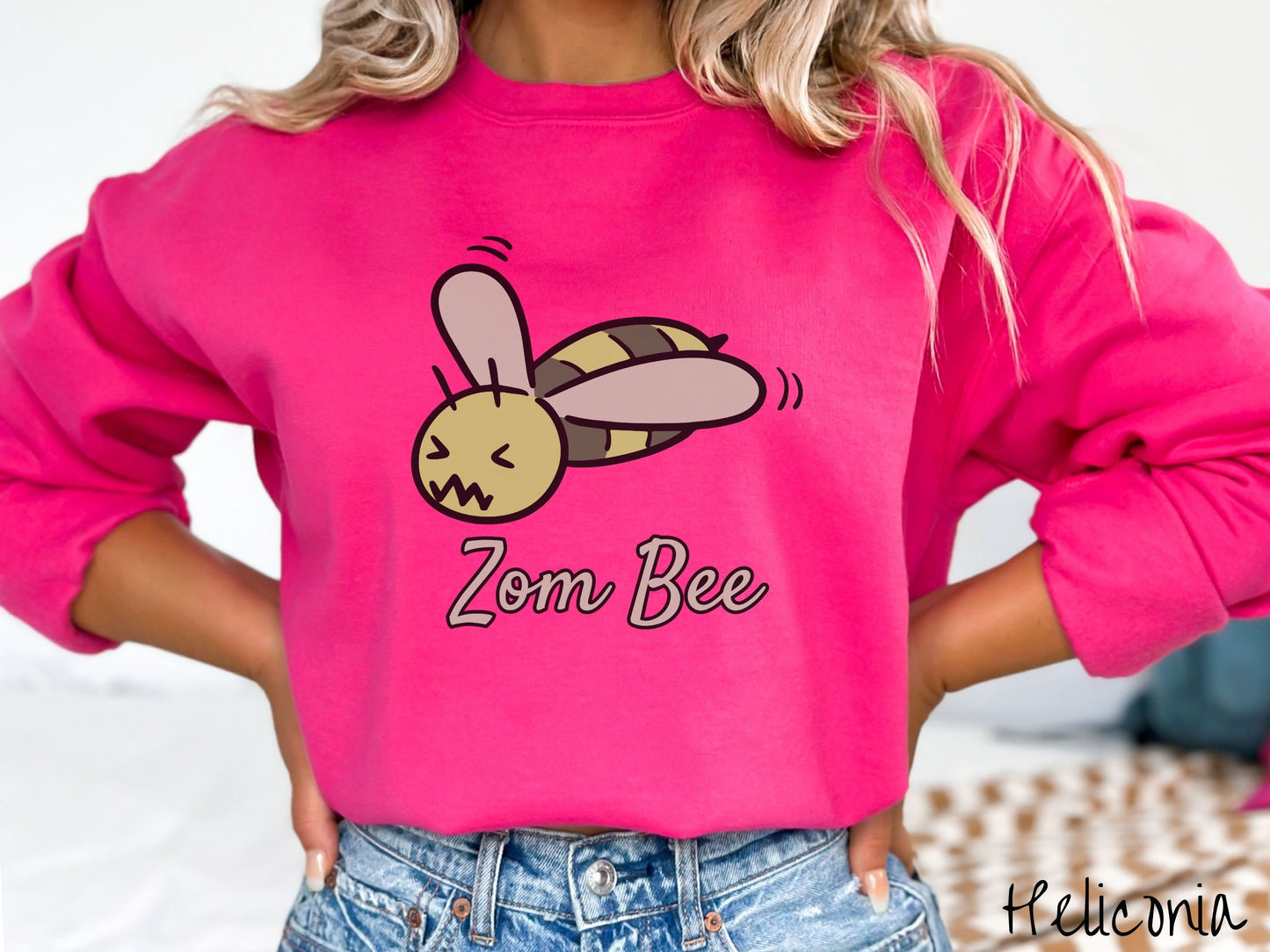 A woman wearing a cute, vintage heliconia colored comfy sweatshirt with the text Zom Bee in light green, cursive font. Above that is a light and dark green honey bee with a crazy face buzzing in the air.