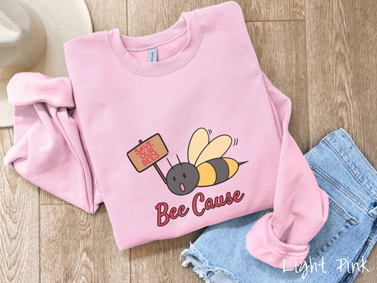 A woman wearing a cute, vintage light pink colored comfy sweatshirt with the text Bee Cause in red, cursive font. Above that is a black and yellow honey bee speaking with its mouth open and holding a sign that reads Save Our Bees in red font.