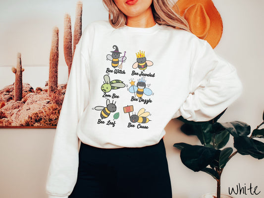 A woman wearing a cute, vintage white colored comfy sweatshirt that has six puns that play on honey bees accompanied with bee drawings. The puns are Bee Witch, Bee Jeweled, Zom Bee, Bee Dazzle, Bee Leaf, and Bee Cause.