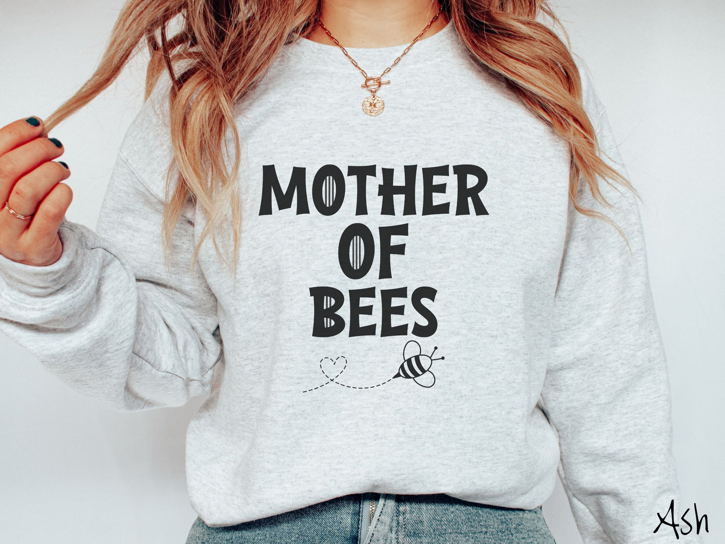 A woman wearing a cute, vintage ash colored comfy sweatshirt with the text Mother of Bees in black, uppercase font. Below that is a black outlined bee buzzing around, the trail it leaves has created a heart shape in the air.