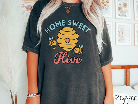 A woman wearing a cute, vintage pepper colored Comfort Colors t-shirt with the text Home Sweet Hive in cyan and red font. In between the words are a yellow bee hive with a red heart on it and smiling honey bees flying around holding flowers.