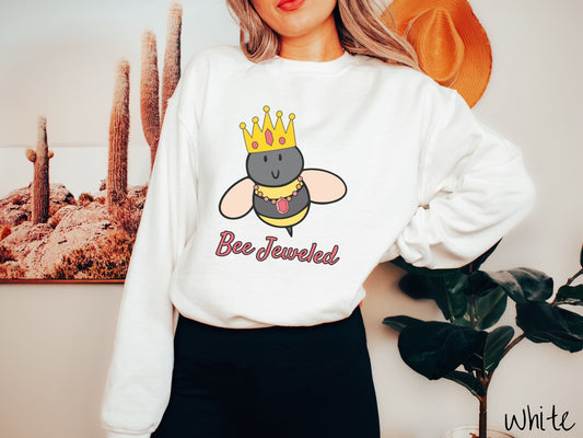 A woman wearing a cute, vintage gold colored comfy sweatshirt with the text Bee Jeweled in pink, cursive font. Above that is a black and yellow honey bee wearing a red, bejeweled golden crown, and a golden necklace with a large, red jewel.