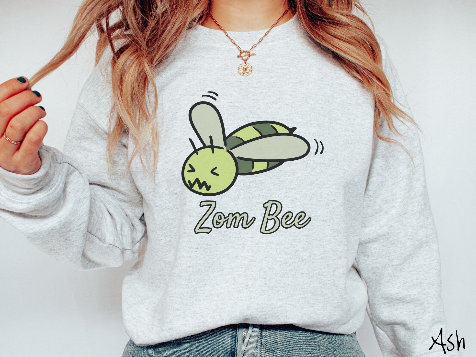 A woman wearing a cute, vintage ash colored comfy sweatshirt with the text Zom Bee in light green, cursive font. Above that is a light and dark green honey bee with a crazy face buzzing in the air.