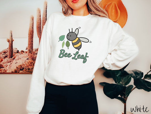 A woman wearing a cute, vintage white colored comfy sweatshirt with the text Bee Leaf in green, cursive font. Above that is a black and yellow honey bee buzzing through the air with green leaves floating next to it.