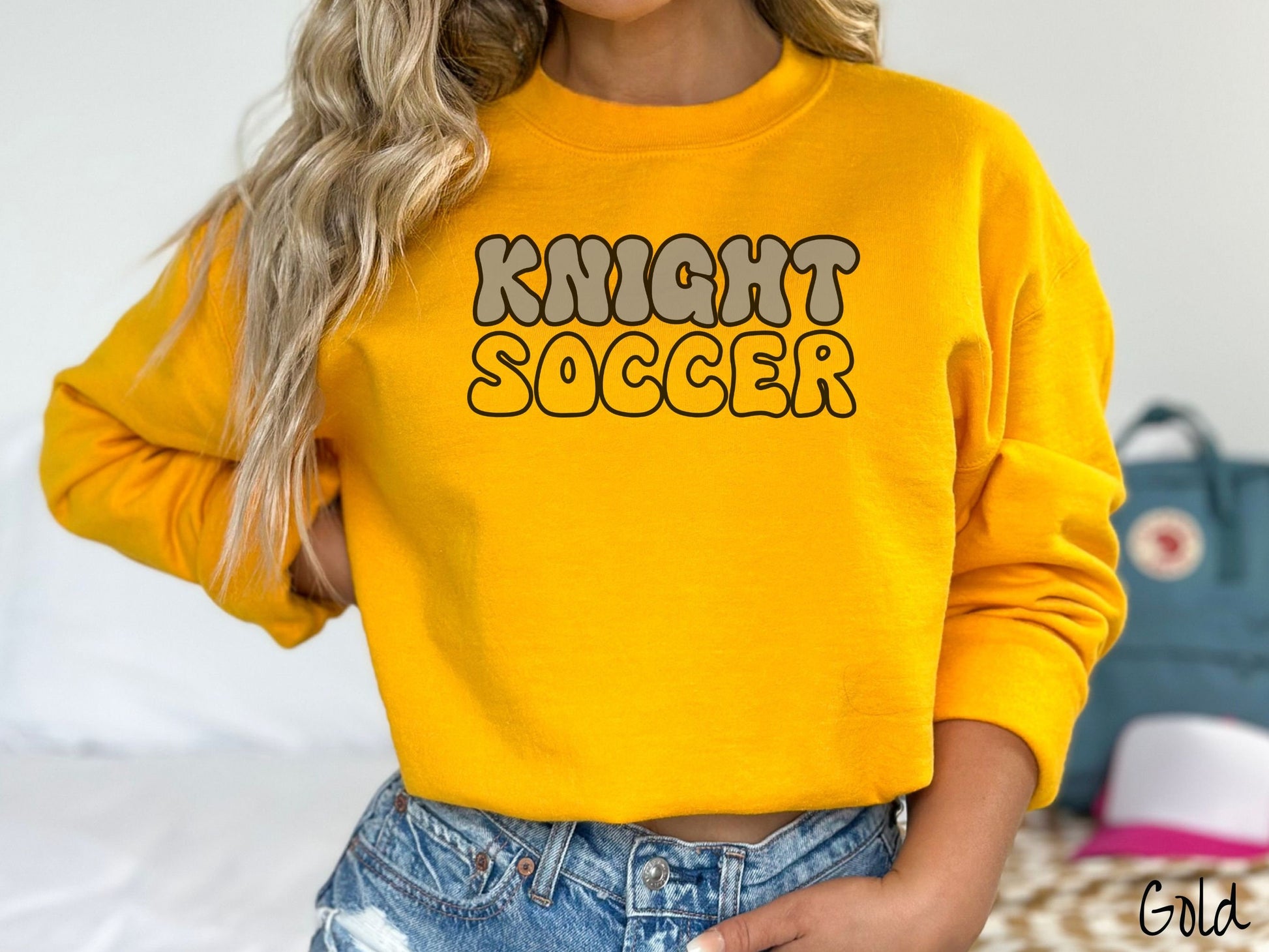 A woman wearing a cute, vintage gold colored sweatshirt with the word Knight across the front in gray, uppercase font. Below that is the word Soccer in gold, uppercase font.