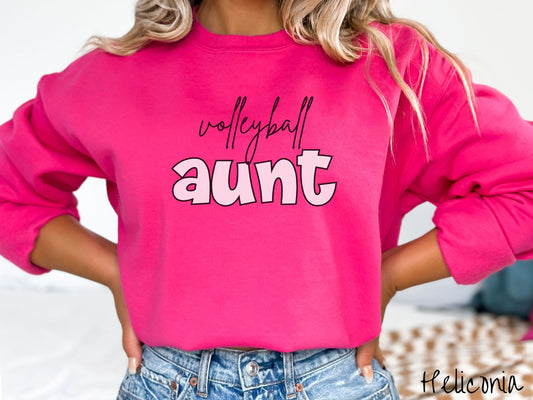 A woman wearing a cute, vintage heliconia colored comfy sweatshirt with the word volleyball across the front in black, undercase cursive writing. Below that is the word aunt in large, light pink font.