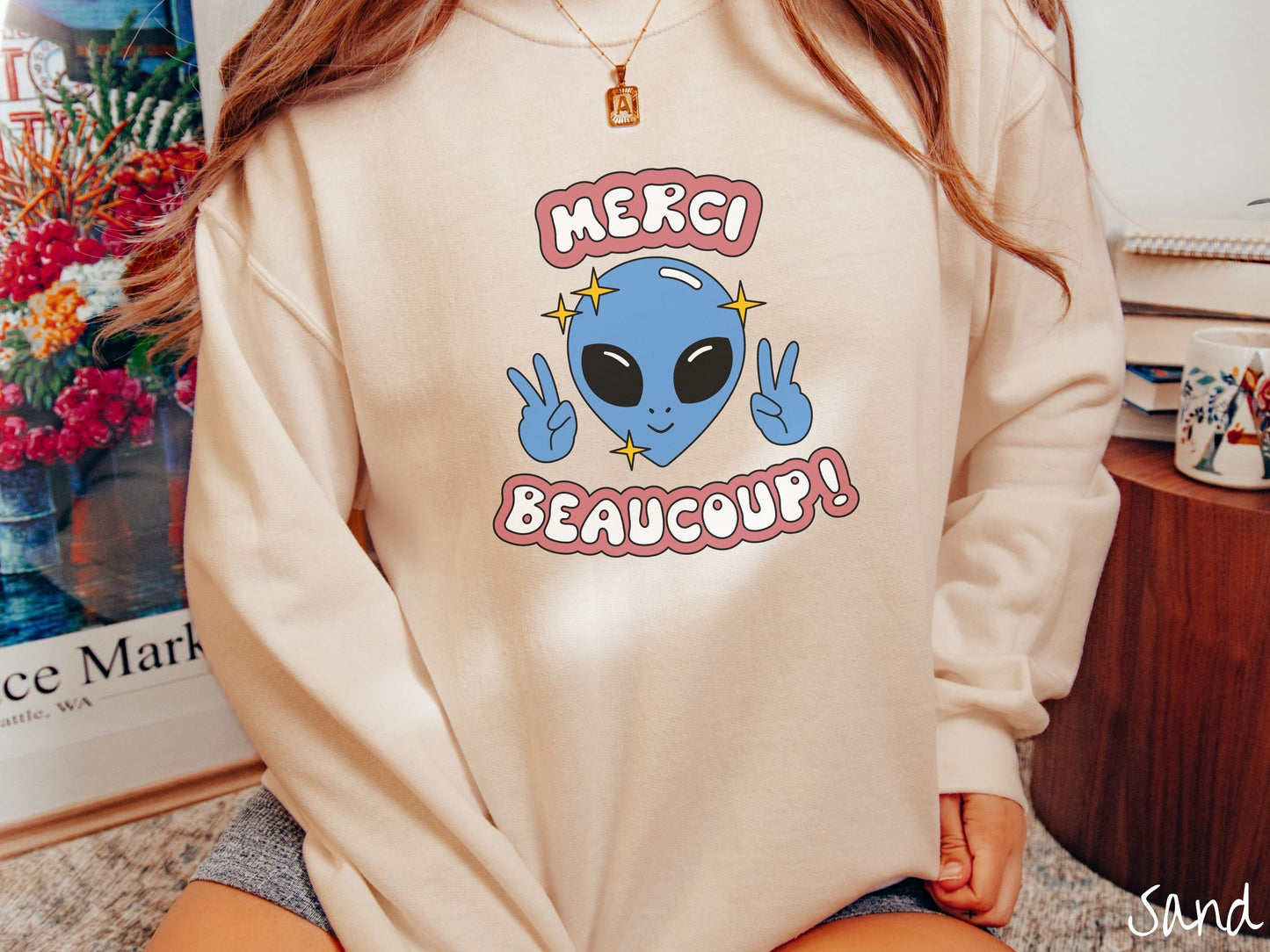 A woman wearing a cute, vintage sand colored comfy sweatshirt with the text Merci Beaucoup! in white and maroon colored font. In between that is a blue alien with large black eyes smiling and holding up two peace signs.