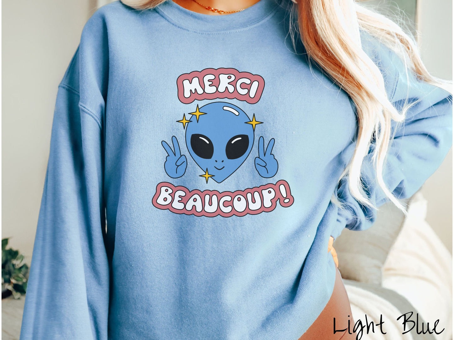 A woman wearing a cute, vintage light blue colored comfy sweatshirt with the text Merci Beaucoup! in white and maroon colored font. In between that is a blue alien with large black eyes smiling and holding up two peace signs.