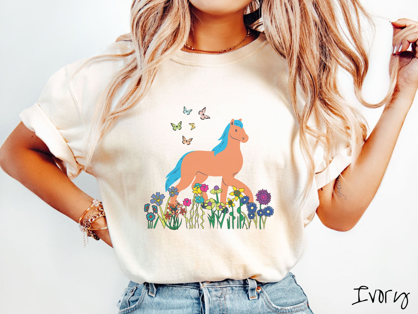 A woman wearing a cute, vintage ivory colored Comfort Colors shirt with a brown horse with blue hair walking through a field of colorful flowers and grass. There are colorful butterflies flying above the horse.