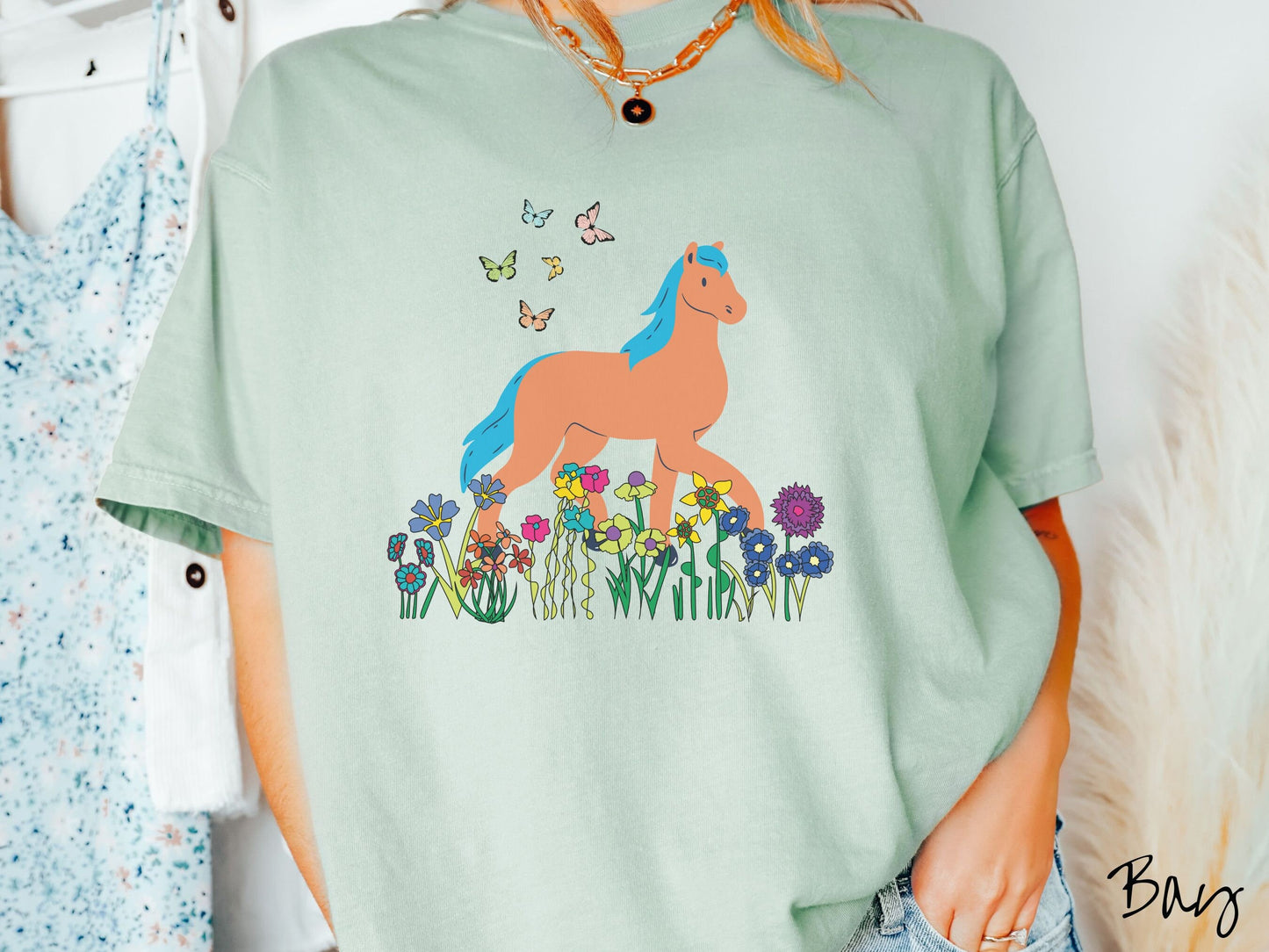A woman wearing a cute, vintage bay colored Comfort Colors shirt with a brown horse with blue hair walking through a field of colorful flowers and grass. There are colorful butterflies flying above the horse.
