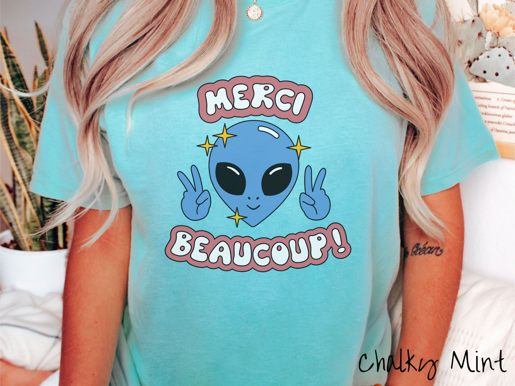 A woman wearing a cute, vintage chalky mint colored Comfort Colors shirt with the text Merci Beaucoup! in white and maroon colored font. In between that is a blue alien with large black eyes smiling and holding up two peace signs.
