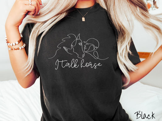 A woman wearing a cute, vintage black colored Comfort Colors shirt with a white silhouette outline of a horse jockey embracing his horse. Below that are the words I talk horse in white, cursive font.