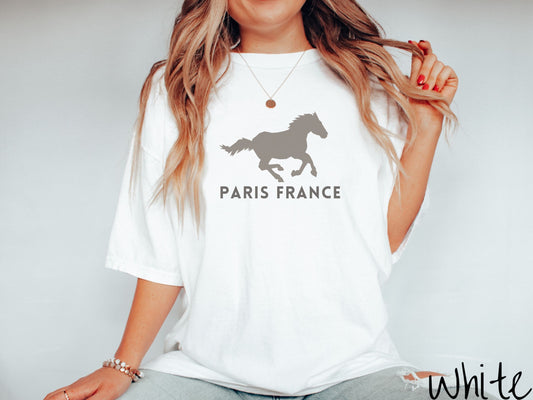 A woman wearing a cute, vintage white colored Comfort Colors shirt with a gray silhouette of a running horse across the font. Below it are the words Paris France in gray, uppercase font.