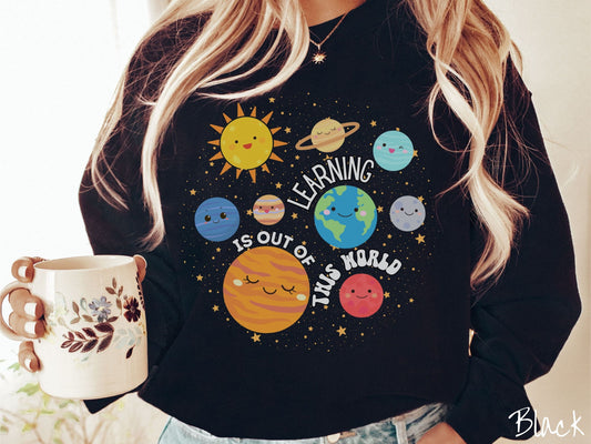 A woman wearing a cute, vintage black colored sweatshirt showing the solar system and the planets have smiling, happy faces. There is text saying Learning Is Out of This World among the planets, and there are many stars twinkling in the background.