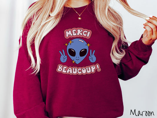 A woman wearing a cute, vintage maroon colored comfy sweatshirt with the text Merci Beaucoup! in white and maroon colored font. In between that is a blue alien with large black eyes smiling and holding up two peace signs.