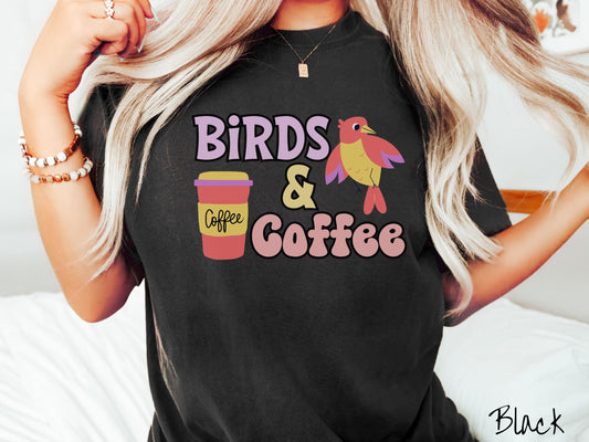 A woman wearing a cute, vintage black colored Comfort Colors shirt with the text Birds & Coffee in purple and light pink font. Next to the text is a red, yellow, and purple smiling bird in the air and a red and yellow to-go coffee cup.