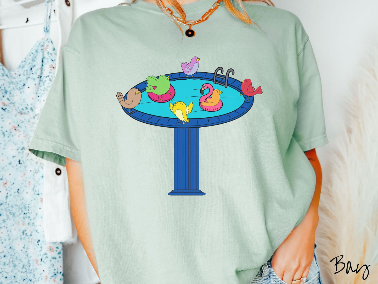 A woman wearing a cute, vintage bay colored Comfort Colors shirt with a blue birdbath with a pool ladder filled with crystal clear blue water. In the water are colorful birds floating on colorful pool inflatables and enjoying the sun.