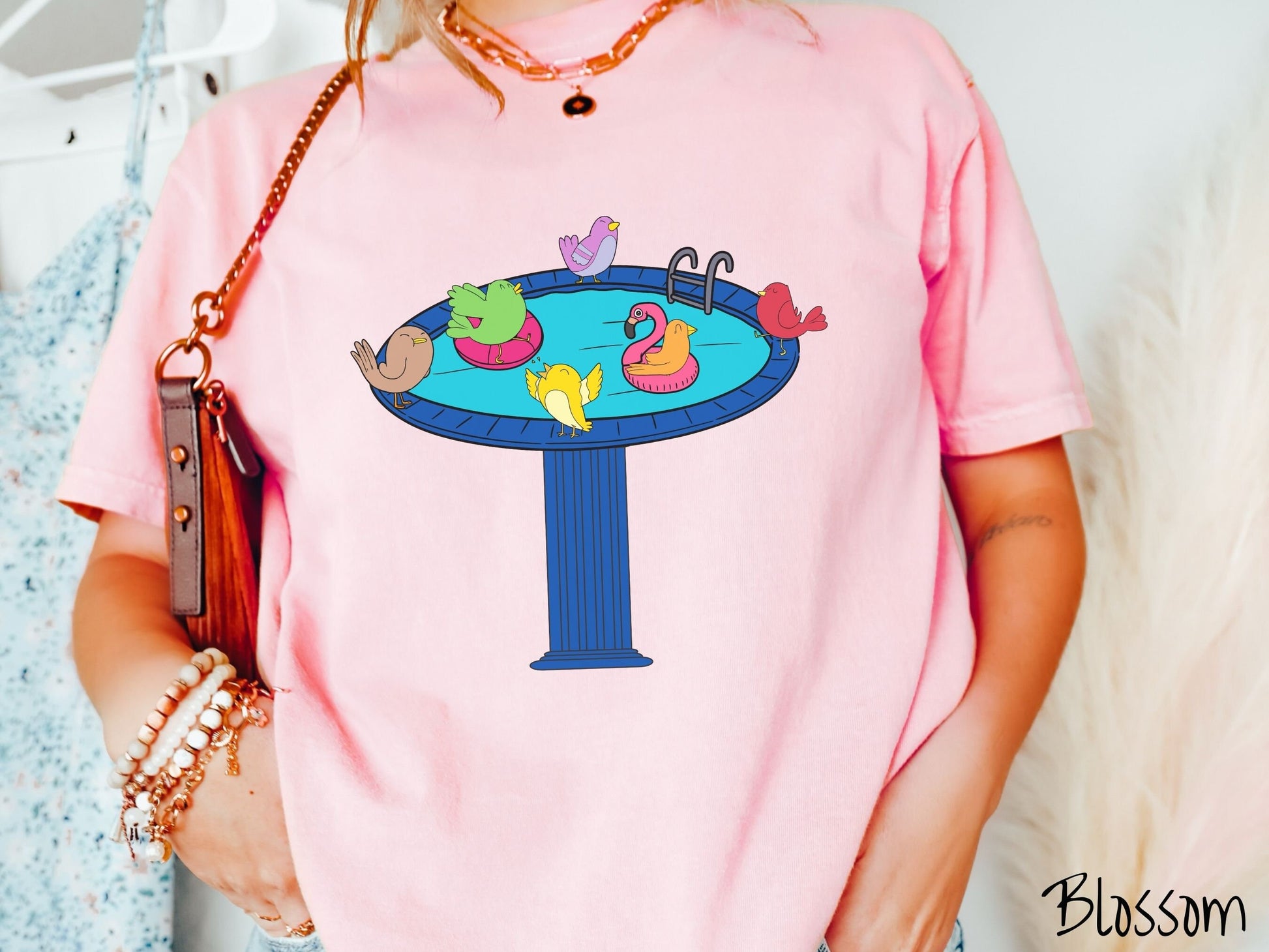 A woman wearing a cute, vintage blossom colored Comfort Colors shirt with a blue birdbath with a pool ladder filled with crystal clear blue water. In the water are colorful birds floating on colorful pool inflatables and enjoying the sun.