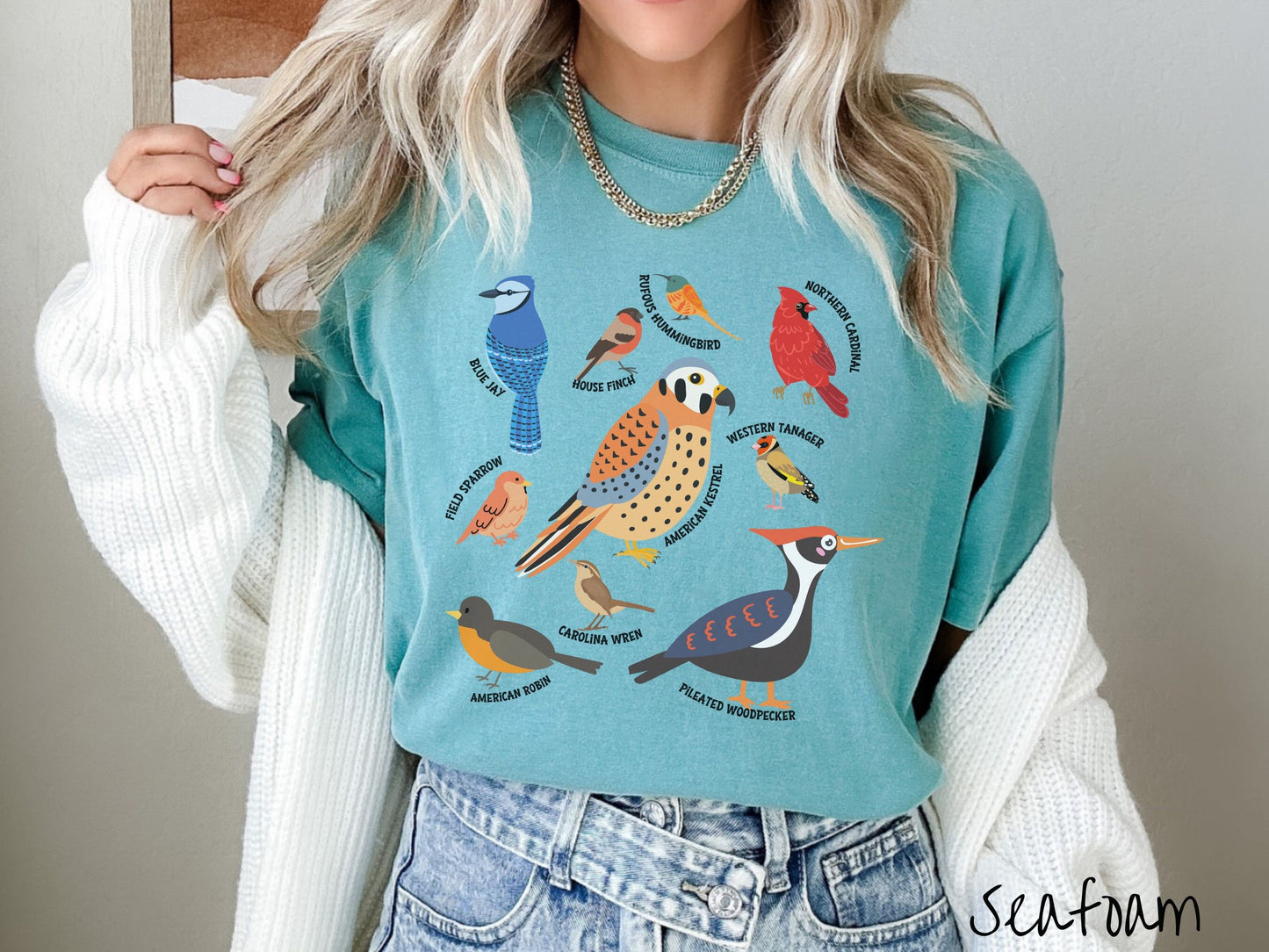 A woman wearing a cute, vintage seafoam colored Comfort Colors shirt with pictures of nine different bird species and their names listed across the front. Some examples are the Blue Jay, House Finch, Western Tanager, and Carolina Wren.
