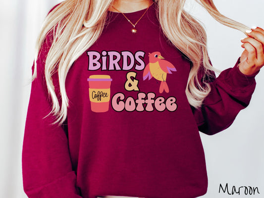 A woman wearing a cute, vintage maroon colored comfy sweatshirt with the text Birds & Coffee in purple and light pink font. Next to the text is a red, yellow, and purple smiling bird in the air and a red and yellow to-go coffee cup.