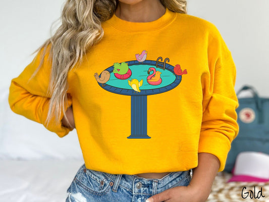 A woman wearing a cute, vintage gold colored comfy sweatshirt with a blue birdbath with a pool ladder filled with crystal clear blue water. In the water are colorful birds floating on colorful pool inflatables and enjoying the sun.