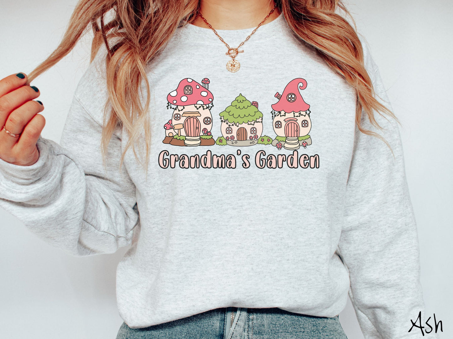A woman wearing a cute, vintage ash colored comfy sweatshirt with the text Grandmas Garden in light pink font. Above the text are cute little cottages for gnomes made from light and dark pink mushrooms.