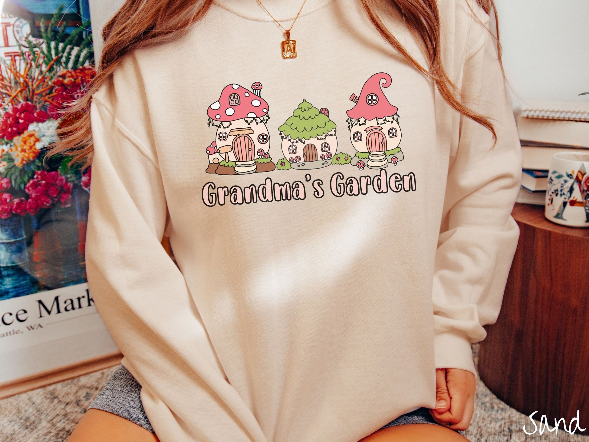 A woman wearing a cute, vintage sand colored comfy sweatshirt with the text Grandmas Garden in light pink font. Above the text are cute little cottages for gnomes made from light and dark pink mushrooms.
