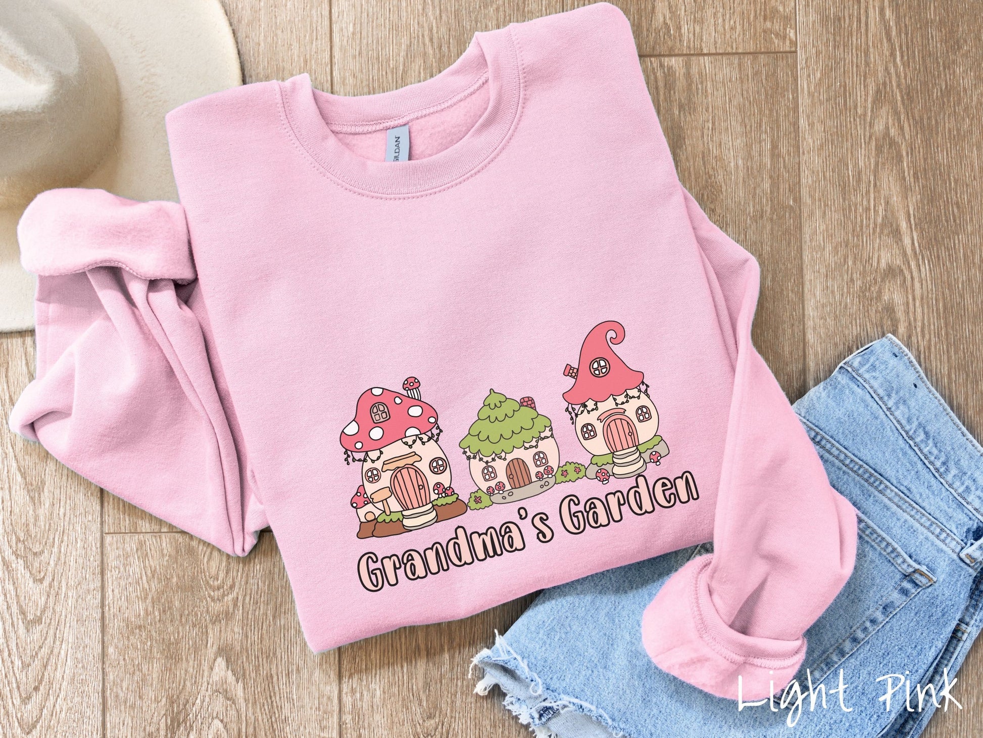 A cute, vintage light pink colored comfy sweatshirt with the text Grandmas Garden in light pink font. Above the text are cute little cottages for gnomes made from light and dark pink mushrooms.