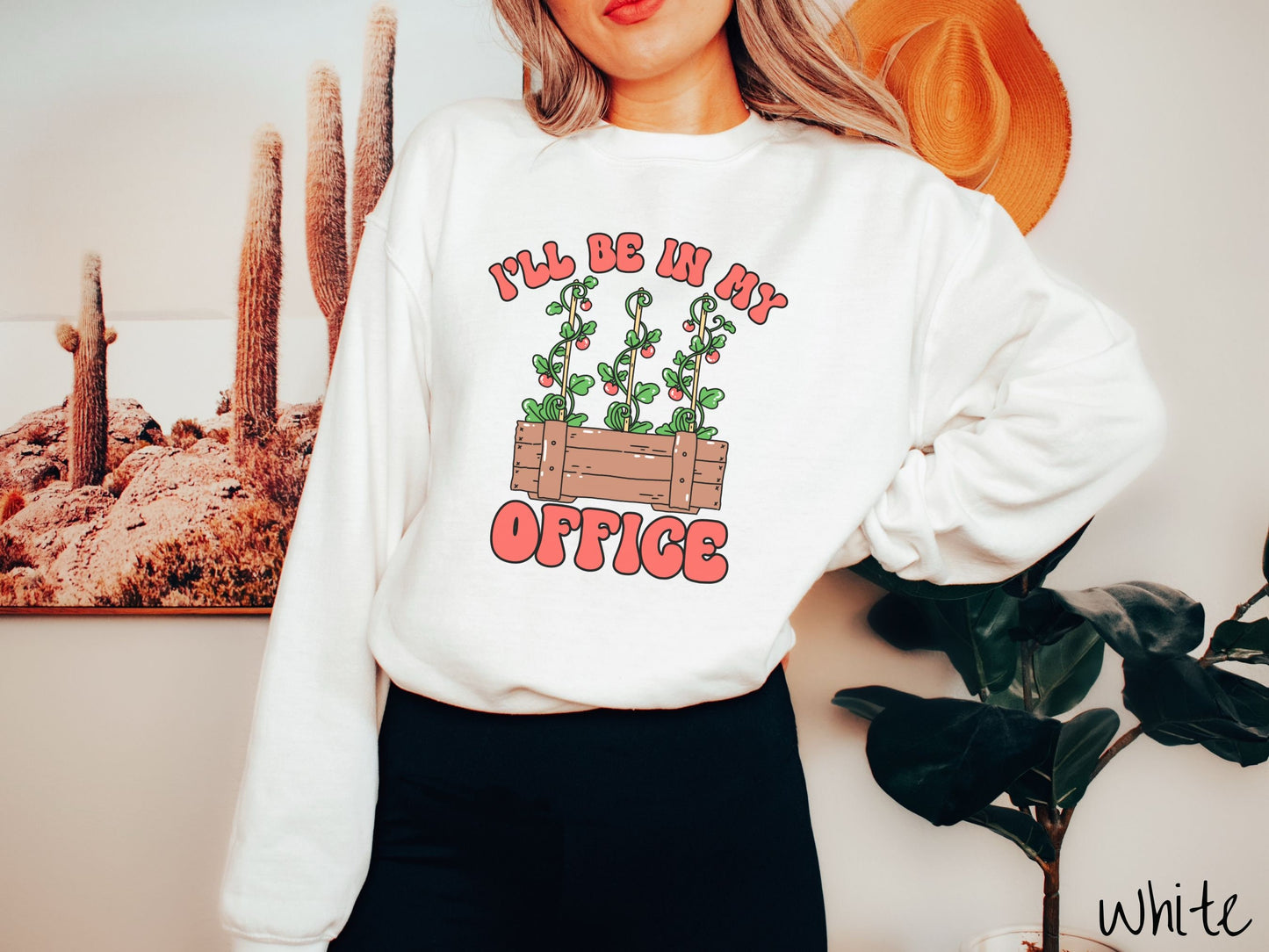 A woman wearing a cute, vintage white colored comfy sweatshirt with the text I’ll Be in My Office in red font across the front. In between the text is a wooden plant holder with three tomato vines sprouting tomatoes climbing upwards.