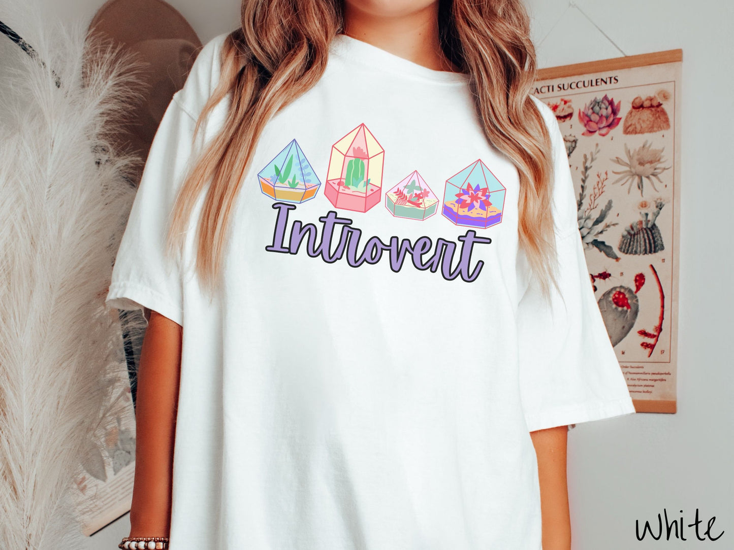 A woman wearing a cute, vintage white colored Comfort Colors t-shirt with the text Introvert in purple font across the front. Above the text are four colorful plant terrariums with colorful flowers and cacti inside.